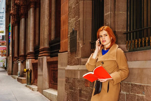 Νεαρή αμερικανική γυναίκα ανάγνωση κόκκινο βιβλίο, μιλάμε για κινητό τηλέφωνο έξω — Φωτογραφία Αρχείου