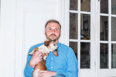 Sakallı, mavi desenli gömlekli genç bir Amerikalı New York 'ta pencere kenarında oturmuş, Dachshund köpeği tutuyor, dinleniyor. En iyi arkadaşı olan genç bir adamın portresi. Köpek.