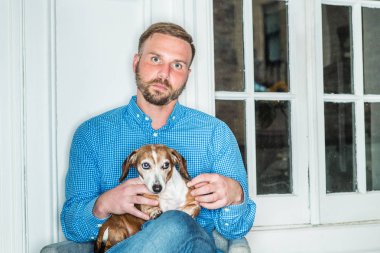 Sakallı, mavi desenli gömlekli genç bir Amerikalı New York 'ta pencere kenarında oturmuş, Dachshund köpeğini kucağında tutuyor, dinleniyor. En iyi arkadaşı olan genç bir adamın portresi. Köpek.