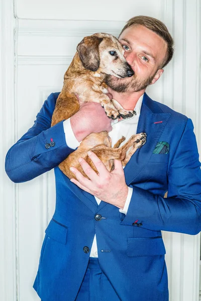 Portrait of young man with best friend - dog. Young American Man with beard, wearing blue suit, standing by white wooden door at home, holding Dachshund dog, kissing. Dog is sick, not feeling well