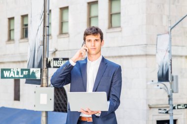 Seyahat eden genç adam, New York 'ta çalışıyor, mavi takım elbiseli, beyaz gömlekli, Wall Street tabelasının yanında ofis binasının önünde duruyor, dizüstü bilgisayarla çalışıyor, cep telefonuyla konuşuyor.