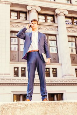 Herhangi bir yeri arıyorum. Seyahat eden genç işadamı, New York 'ta çalışıyor, mavi takım elbise giyiyor, beyaz gömlek giyiyor, dizüstü bilgisayarı tutuyor, ofis binasının önünde duruyor, cep telefonuyla konuşuyor.