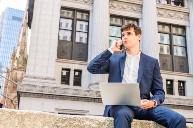 Teknolojinin gücü. Seyahat eden, New York 'ta çalışan, mavi takım elbiseli, beyaz gömlekli, ofis binasının önünde oturan, dizüstü bilgisayarda çalışan, cep telefonuyla konuşan genç bir işadamı.