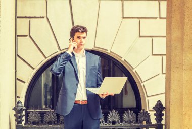 Teknolojinin gücü. New York 'ta uzaktan çalışan genç bir işadamı, mavi takım elbiseli, beyaz gömlekli, ofisin önünde duran, dizüstü bilgisayarla çalışan, cep telefonuyla konuşan