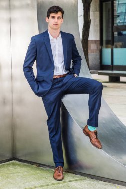 New York 'taki Genç Yakışıklı İş Adamı portresi. Mavi takım elbiseli genç bir üniversite öğrencisi, beyaz gömlek, kahverengi deri ayakkabılar, elleri ceplerinde, gümüş metal duvara yaslanmış,