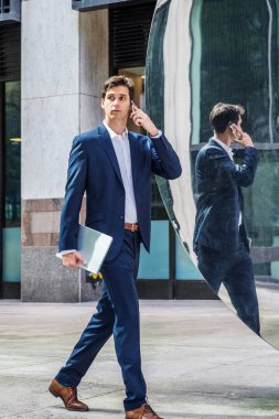Seyahat eden, New York 'ta çalışan, mavi takım elbiseli, beyaz tişörtlü, dizüstü bilgisayarlı, metal aynanın önünden geçen, cep telefonuyla konuşan genç bir işadamı.