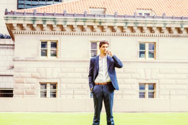 Modern günlük yaşam. New York 'ta çalışan yakışıklı genç işadamı, mavi takım elbiseli, beyaz gömlekli, el ele tutuşmuş, yeşil çimlerin üzerinde ofis binasının önünde duruyor, cep telefonuyla konuşuyor.