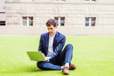 Genç üniversite öğrencisi, New York 'ta çalışıyor, mavi takım elbise giyiyor, beyaz gömlek giyiyor, kahverengi deri ayakkabılar giyiyor, kampüsün dışındaki yeşil çimlerde oturuyor, dizüstü bilgisayarla çalışıyor.