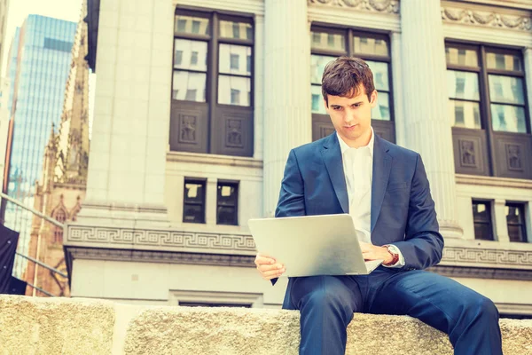 Modern daily life. Young businessman traveling, working in New York City, wearing blue suit, white shirt, sitting on street outside office building, looking down, working on laptop computer