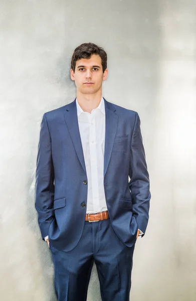 Portrait of Young Handsome Businessman in New York City. Young college student wearing blue suit, white shirt, hands in pockets, standing against silver metal background, looking forward, thinking