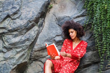 Genç melez Afro kıvırcık saçlı, New York, Central Park 'ta kırmızı kitap okuyan, kırmızı çiçekli desenli elbisesi olan yeşil yapraklı kayaların üzerinde oturan Afrikalı Amerikalı kadın.