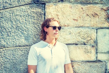 New York 'ta uzun koyu kahverengi saçlı, beyaz kısa kollu gömlek, güneş gözlüğü takan, yaz aylarında güneş altında kayaların karşısında duran, uzun boylu, yakışıklı bir Amerikalı.