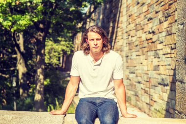 New York 'ta uzun koyu kahverengi saçlı, beyaz kısa kollu gömlekli, mavi kotlu, yaz aylarında güneşin altında yarı duvarda oturan genç, yakışıklı Amerikalı adamın portresi.