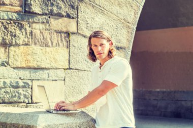 New York 'un dışında dizüstü bilgisayarda çalışan, beyaz kısa kollu tişörtlü, uzun siyah saçlı genç Amerikalı adam, kampüsün kayalık kapısında güneşin altında duruyor, başka yöne bakıyor, düşünüyor.