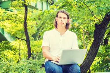 New York 'un dışında dizüstü bilgisayarla çalışan uzun koyu kahverengi saçlı genç Amerikalı adam, beyaz kısa kollu gömlek giyiyor, yeşil ağaçların arkasında oturuyor, başka tarafa bakıyor ve düşünüyor.