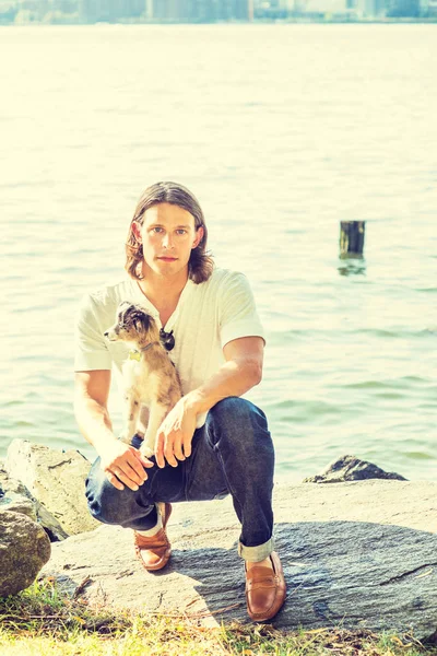 Portrait of man with best friend - dog. Young American Man with long hair, wearing white shirt, jeans, brown leather shoe, squatting on rocks by Hudson River  in New York City, with dog on lap, relax