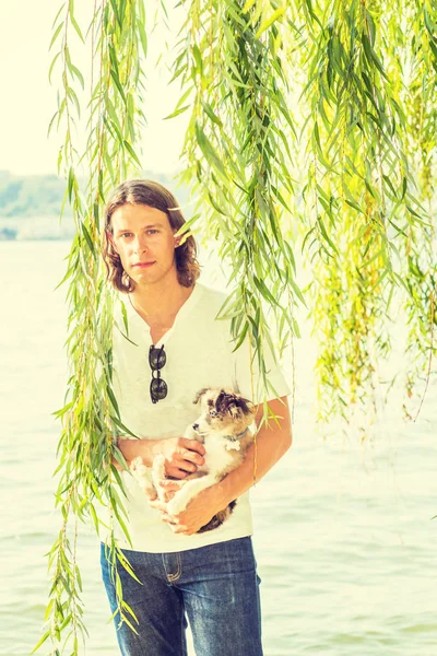 Portrait of man with best friend - dog. Young American Man with long hair, wearing white shirt, jeans, carrying dog with arms, standing by Hudson River with green willow leaves in New York City, rela