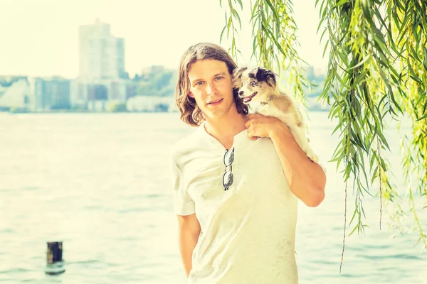 Portrait of man with best friend - dog. Young American Man with long hair, wearing white shirt, carrying dog on shoulder, standing by Hudson River with green willow leaves in New York City, relaxing