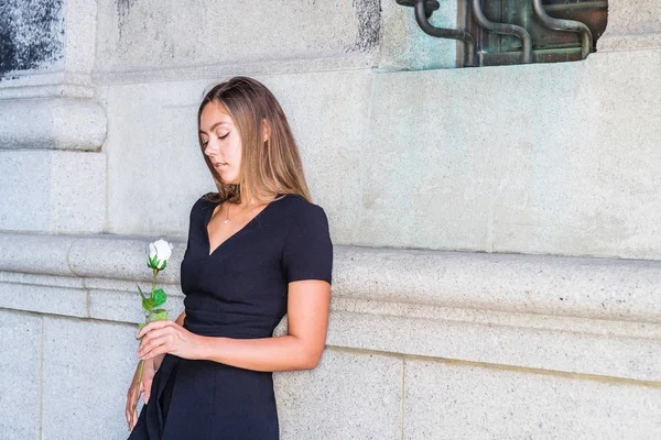 I love you, waiting for you. Young East European Woman wearing black short sleeve, v neck dress, holding white rose flower, standing by wall on street in New York City, looking down, sad, thinking