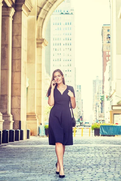 若い東ヨーロッパの女性は長い髪を持ち 携帯電話で話し ニューヨークを旅し 黒い半袖 Vネックドレス ハイヒールを着て ショルダーバッグを持って ヴィンテージストリートを歩く — ストック写真