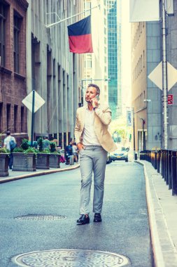 Genç Avrupalı iş adamı seyahat ediyor, New York 'ta çalışıyor, sakallı, gri saçlı, bej ceketli, gri pantolonlu, siyah deri ayakkabılı, dar bir sokakta yürüyor, cep telefonuyla konuşuyor.