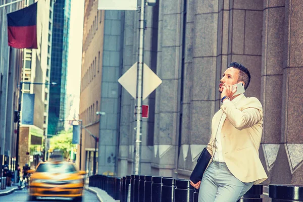 Genç Avrupalı iş adamı seyahat ediyor, New York 'ta çalışıyor, sakallı, gri saçlı, bej ceketli, gri pantolonlu, evrak çantalı, eski sokakta oturuyor, cep telefonuyla konuşuyor.
