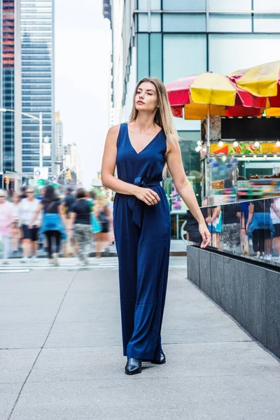 Young Woman Street Fashion. Young Eastern European Woman traveling in New York City, wearing blue sleeveless jumpsuit, black leather shoes, walking on busy street in middletown of Manhattan, relaxing.