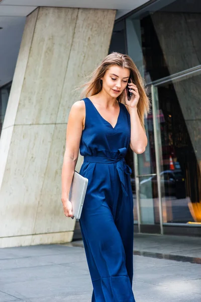 年轻的东欧女人 会讲手机 在纽约工作 穿蓝色无袖连衣裙 带着手提电脑 走在办公楼外的街上 — 图库照片