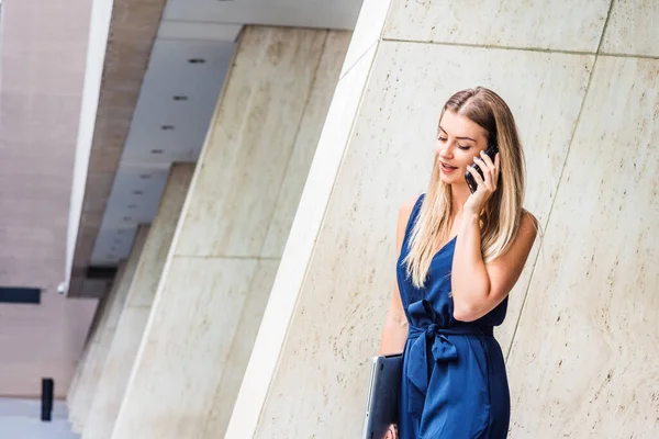 Young Eastern European American Woman talking on phone, traveling, working in New York City, wearing blue jumpsuit, carrying laptop computer, looking down, walking out from office building to street