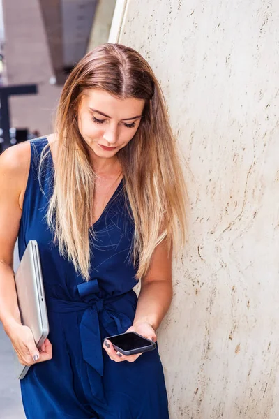 長い茶色の髪をした若い東欧系アメリカ人女性 青い袖のないジャンプスーツを着て ラップトップコンピュータを運ぶ ニューヨーク市のオフィスの外に立って 携帯電話でテキストメッセージ — ストック写真
