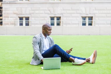 New York 'ta çalışan, gri desenli spor ceket, mavi pantolon, spor ayakkabı giyen, yeşil çimlerde oturan, dizüstü bilgisayarda çalışan, cep telefonuyla mesajlaşan genç Afrikalı Amerikalı iş adamı.