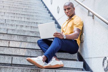 New York 'ta çalışan, sarı gömlekli, mavi pantolonlu, beyaz spor ayakkabılı, ofis binasının dışındaki merdivenlerde oturan, dizüstü bilgisayarında çalışan, yüzünü çevirmiş,