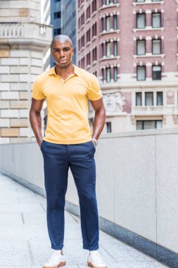 New York 'ta genç, Afro-Amerikan erkek günlük moda, sarı kısa kollu gömlek, mavi pantolon, beyaz spor ayakkabı, elleri cepte, kampüsün dışında duruyor, ileriye bakıyor.