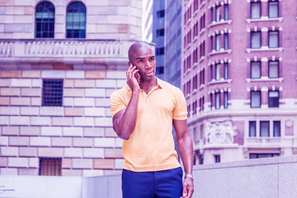 Young African American college student with short hair, wearing yellow short sleeve shirt, standing outside office building on campus in New York City, talking on cell phone. Color filtered effect