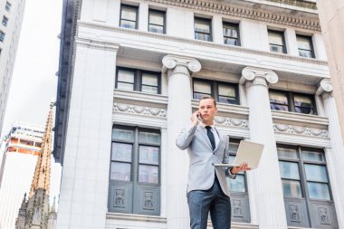 Seyahat eden genç iş adamı, New York 'ta çalışıyor, gri ceket, beyaz gömlek, siyah kravat giyiyor, klasik ofis binasının önünde duruyor, dizüstü bilgisayarla çalışıyor, cep telefonuyla konuşuyor.