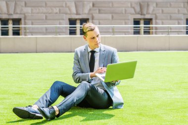 Genç Avrupalı Adam çalışıyor, New York 'ta çalışıyor, gri ceket, beyaz gömlek, siyah kravat, siyah pantolon, deri ayakkabı, dizüstü bilgisayar, yeşil çimlerin üzerinde oturuyor, güneş altında,