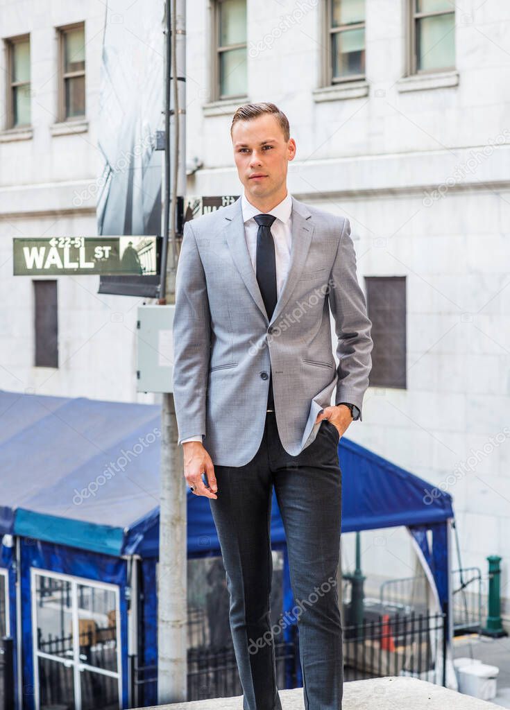 Young European Businessman traveling in New York City, wearing gray blazer, white undershirt, black tie, black pants, standing on street outside office building by Wall Street sign, looking forward