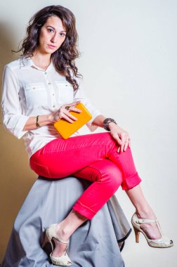 Beyaz uzun kollu gömlek, kırmızı pantolon, açık renkli sandaletler giyen, küçük sarı deri çanta tutan, sana bakan güzel bir kadın. Stüdyo çekimi