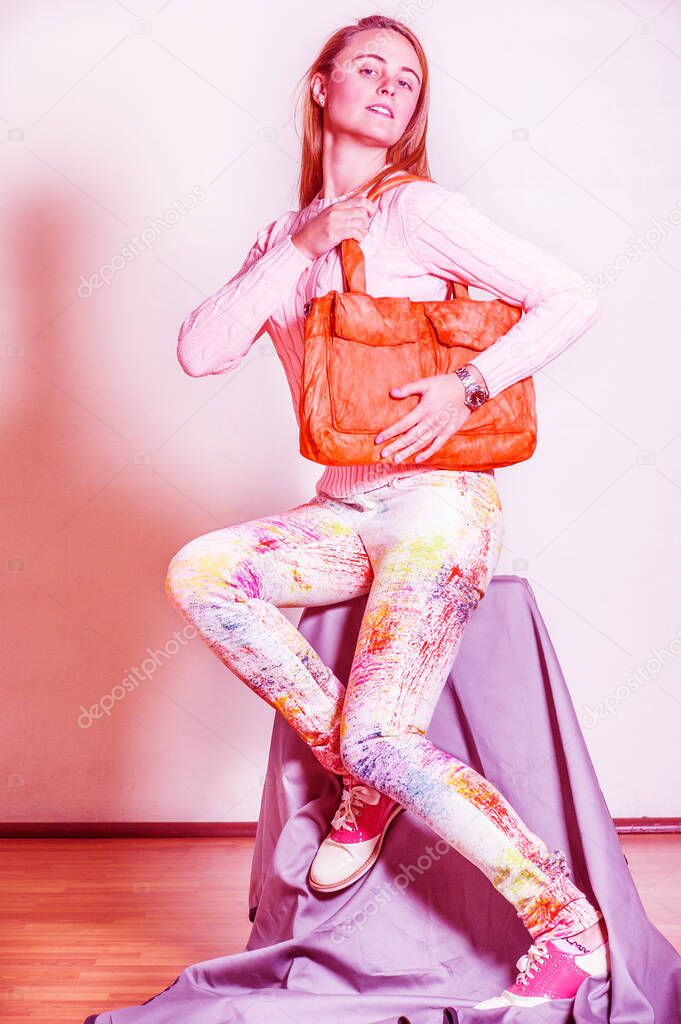 Young woman wearing long sleeve light pink knit sweater, light colorful pants, red and white leather sneakers, shoulder carrying orange brown leather bag. Color filtered effec