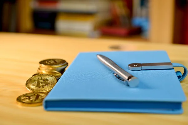 Sobre a mesa de ouro criptomoeda moedas, caderno azul com caneta, prateleira borrada com livros no fundo — Fotografia de Stock