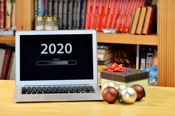 Op tafel kerstversiering, geschenkdoos en laptop met tekst - 2020 laden - op het scherm, wazig schap met boeken op de achtergrond — Stockfoto