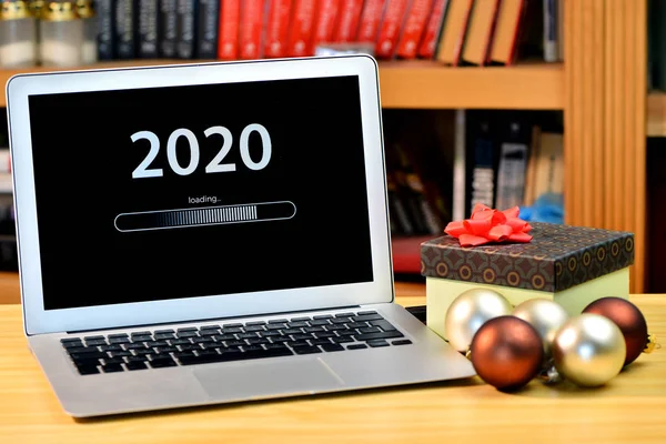 Op tafel kerstversiering, geschenkdoos en laptop met tekst - 2020 laden - op het scherm, wazig schap met boeken op de achtergrond — Stockfoto