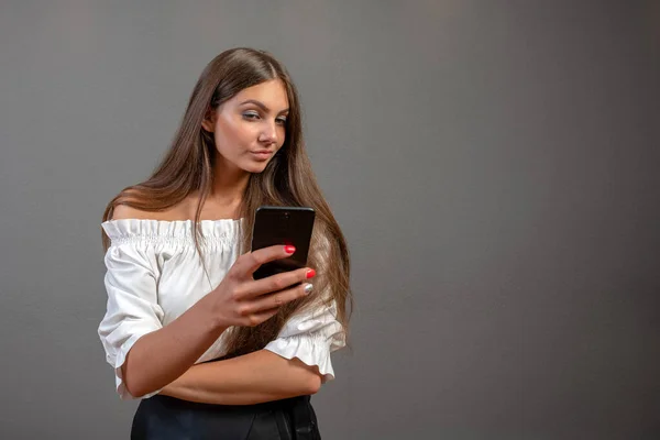 Photo d'une femme souriante aux longs cheveux foncés tenant et utilisant un téléphone portable noir isolé sur fond gris — Photo