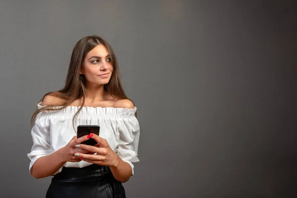 Foto einer lächelnden Frau mit langen dunklen Haaren, die ihr schwarzes Mobiltelefon über grauem Hintergrund isoliert hält — Stockfoto