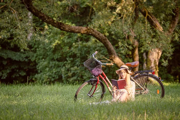 Una giovane, magra, bionda donna nel parco siede accanto alla sua moto e legge un libro. Autunno tonalità di colore — Foto Stock