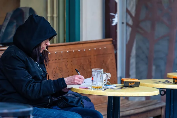 Ginebra, Suiza - 14 de abril de 2019: Un hombre con una capucha en la cabeza está sentado en una mesa en un café al aire libre, fumando y escribiendo un documento —  Fotos de Stock