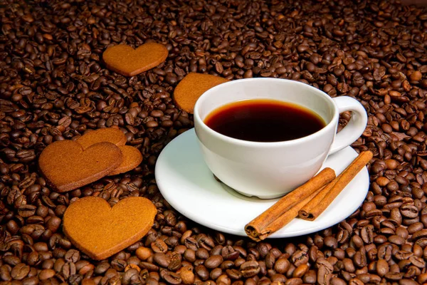 Taza de café blanco, palitos de canela y galletas de jengibre en forma de corazón en el fondo de granos de café — Foto de Stock