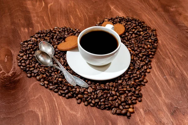 Caneca de café branca, pão de gengibre em forma de coração e duas colheres em uma base em forma de coração feita de grãos de café — Fotografia de Stock