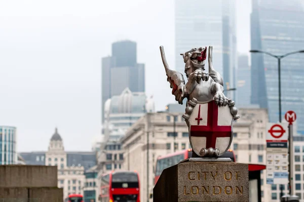 Uma escultura de dragão com o Brasão de Armas da Cidade de Londres na Ponte de Londres. Arranha-céus desfocados e autocarros vermelhos de dois andares no fundo — Fotografia de Stock