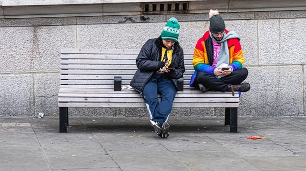 Двое мужчин сидят на скамейке на обочине улицы и смотрят на свои смартфоны — стоковое фото